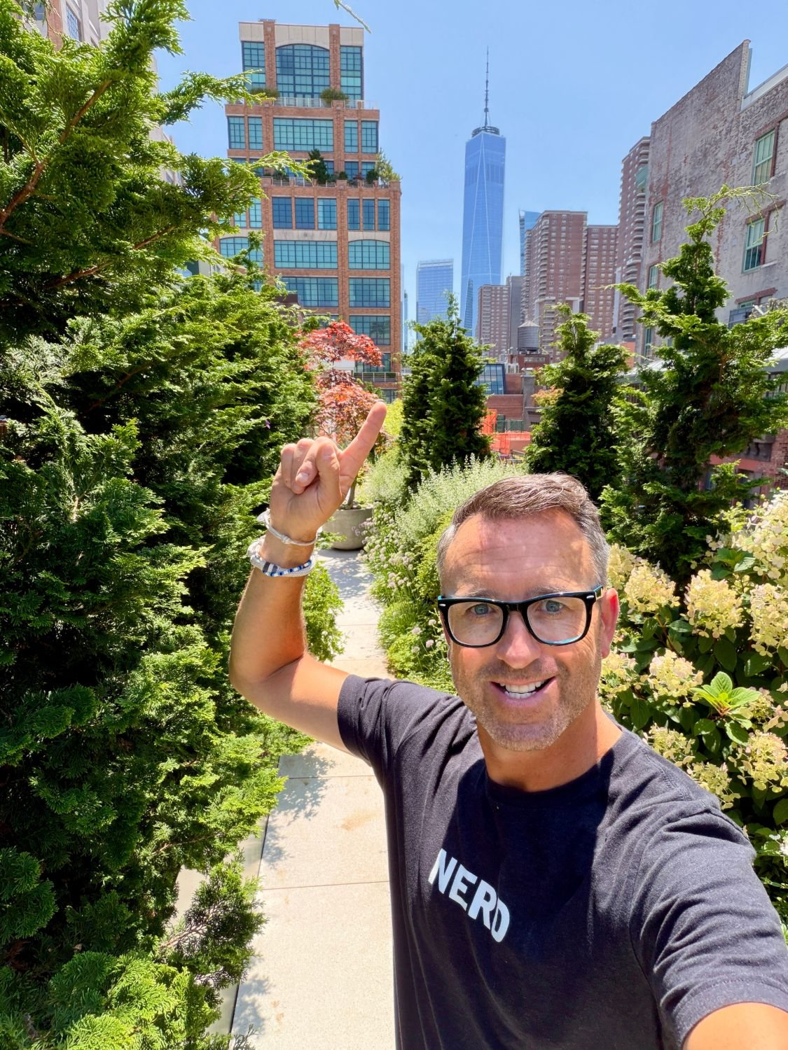 Andy Humphrey in a NERD t-shirt on a Manhattan rooftop garden, pointing up, with One World Trade Center behind him